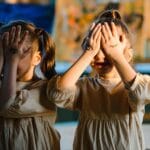 Dwie bliźniaczki bawią się w akuku w domu, zasłaniając twarze pomalowanymi dłońm