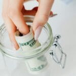 Dłoń wkłada zwinięte banknoty dolarowe do szklanego słoika z oszczędnościami
