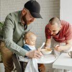 Dwóch ojców karmi w domu uśmiechniętego malucha przy stole