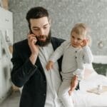 Tata z brodą trzyma niemowlę na rękach i rozmawia przez telefon